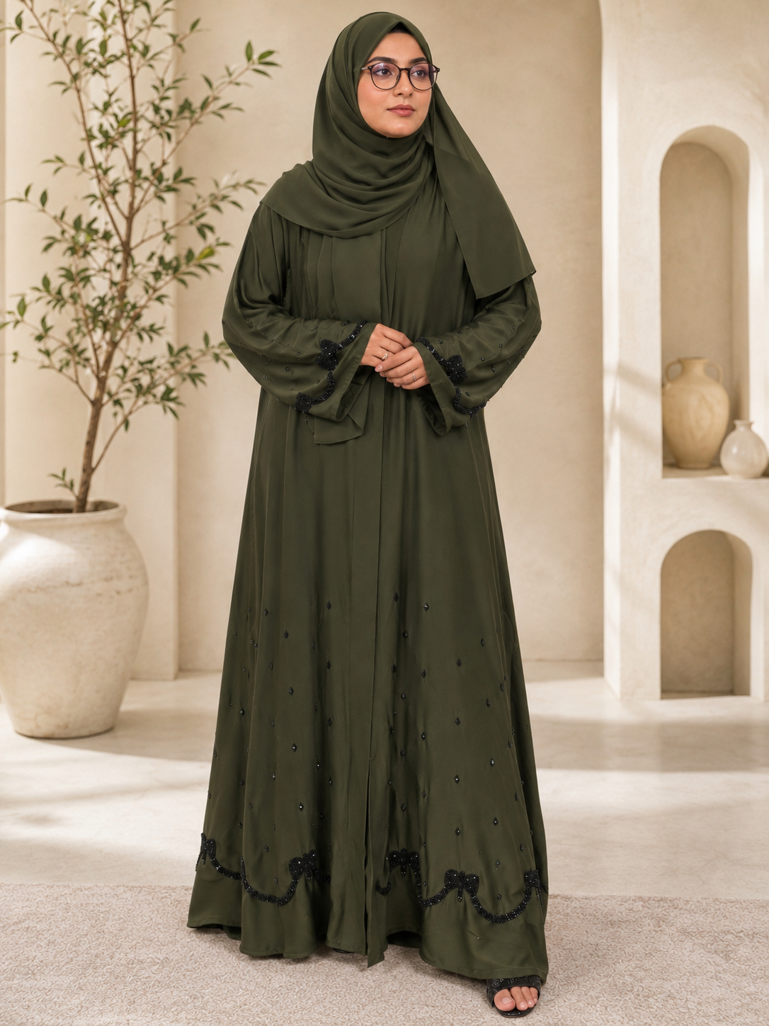 Full-length studio portrait of a model wearing the Olive Arcana abaya in premium imported olive green Nidha fabric, featuring a flowing silhouette, hand-beaded black glass scalloped trim on the hem and cuffs, scattered crystal accents, and a matching olive hijab in a luxury archway setting