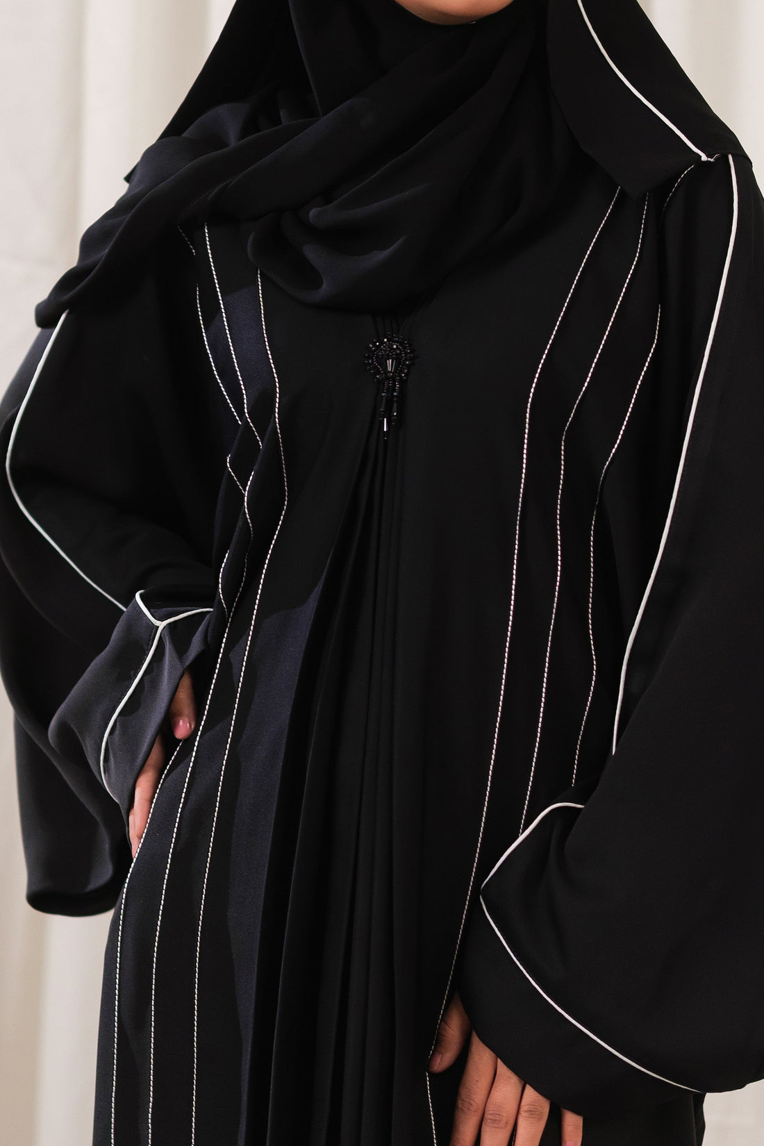 Dhaagey Meridian Abaya in black, showing the voluminous silhouette, central pleated design, curved CTC paneling with White Piping, and the black beaded ornament.