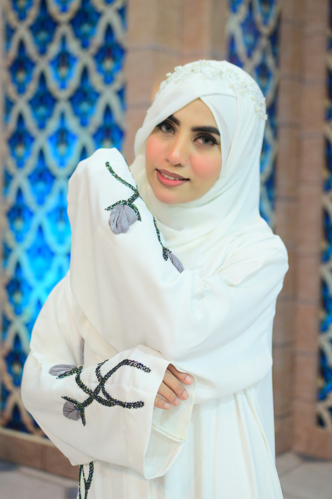 A premium ivory white Nidha abaya featuring a vertical hand-embellished 3D floral vine with grey fabric petals and beadwork down the front, modeled in a luxury setting.