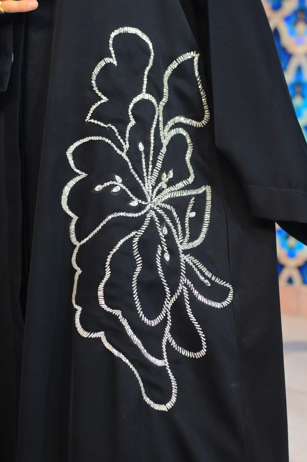 A premium black Nidha abaya featuring an oversized, hand-embellished silver floral motif with intricate beadwork on the side and sleeve, modeled in a luxury studio setting.