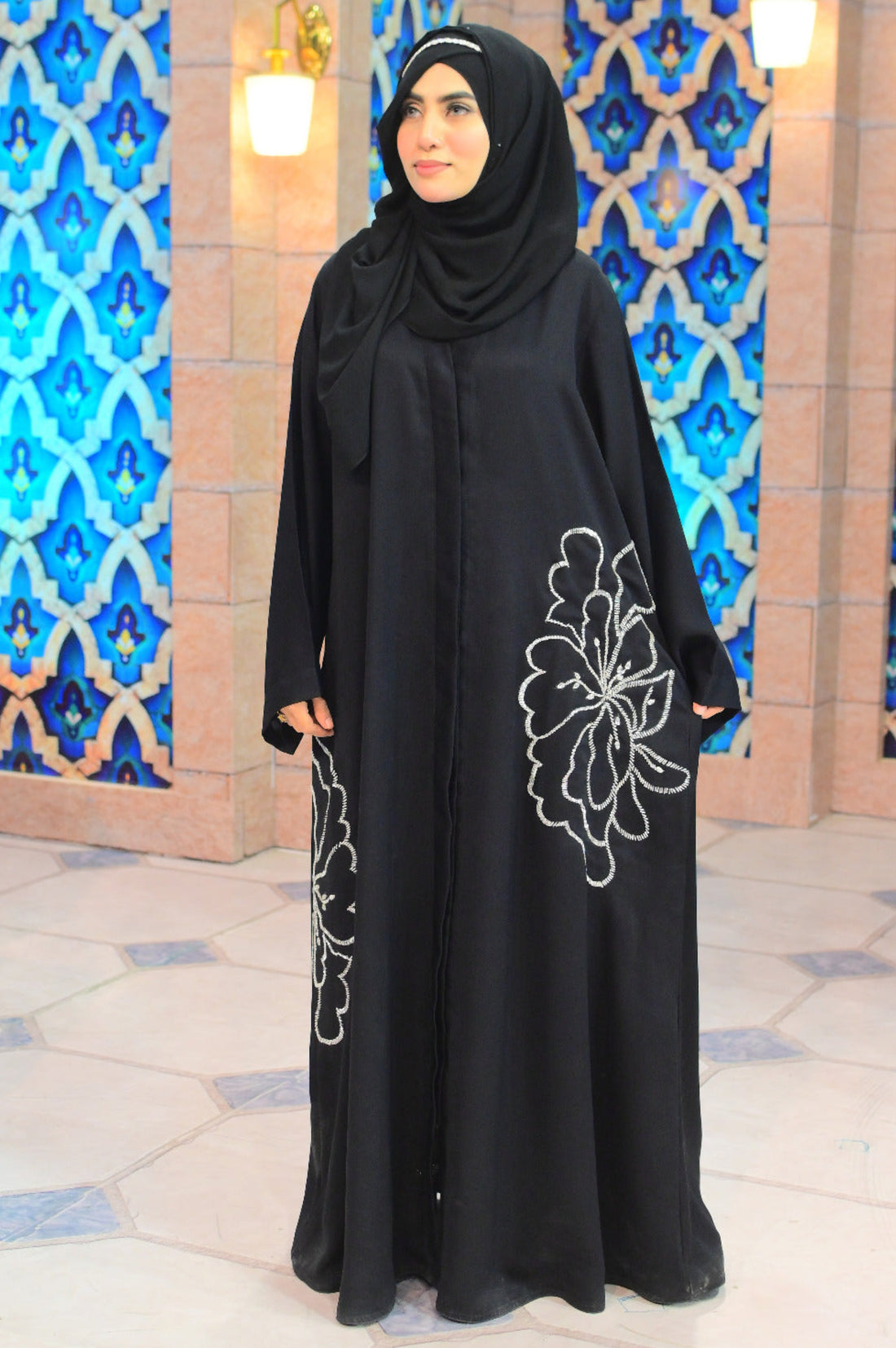 A premium black Nidha abaya featuring an oversized, hand-embellished silver floral motif with intricate beadwork on the side and sleeve, modeled in a luxury studio setting.